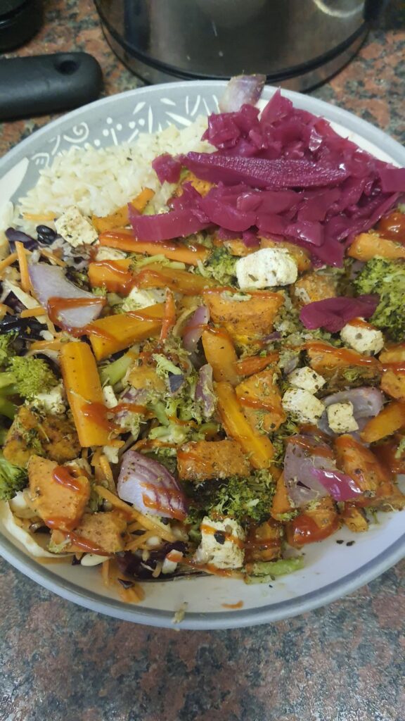 Meal photo, roasted vegetables in a bowl with rice and pickled cabbage. Vegetables included are carrots, sweet potatoe, broccoli, red onion along with tofu. 